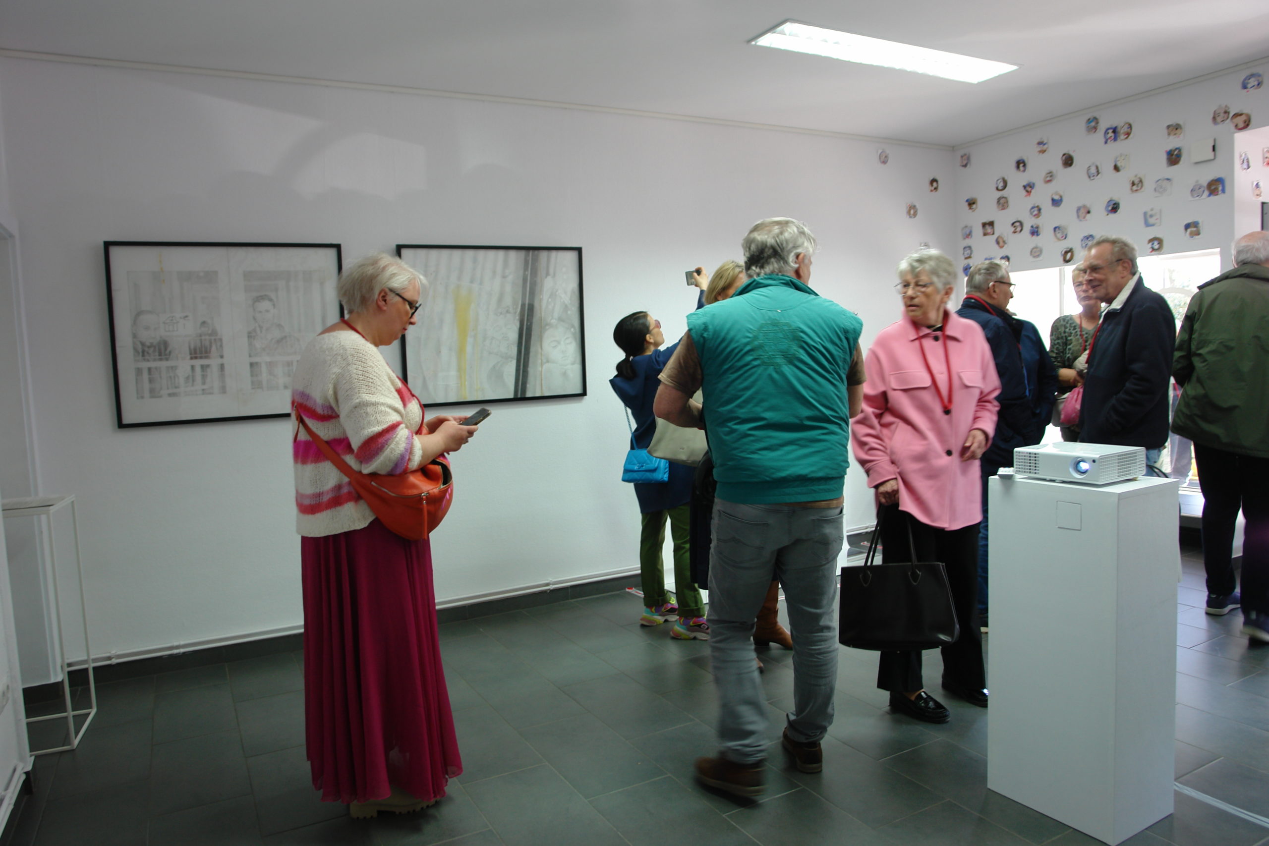 Galerieraum: Einzelne zeichnerische Arbeiten sind an den Wänden zu sehen. Davor stehen Menschen und schauen sich diese an oder unterhalten sich.